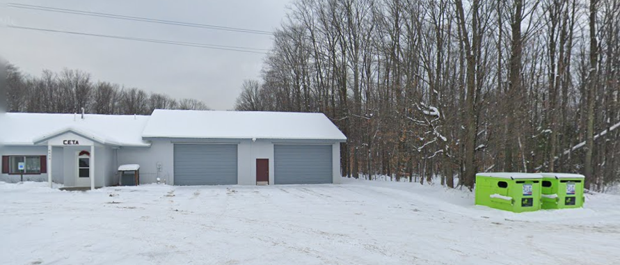 Snow-covered building near forest, green recycling bins nearby.